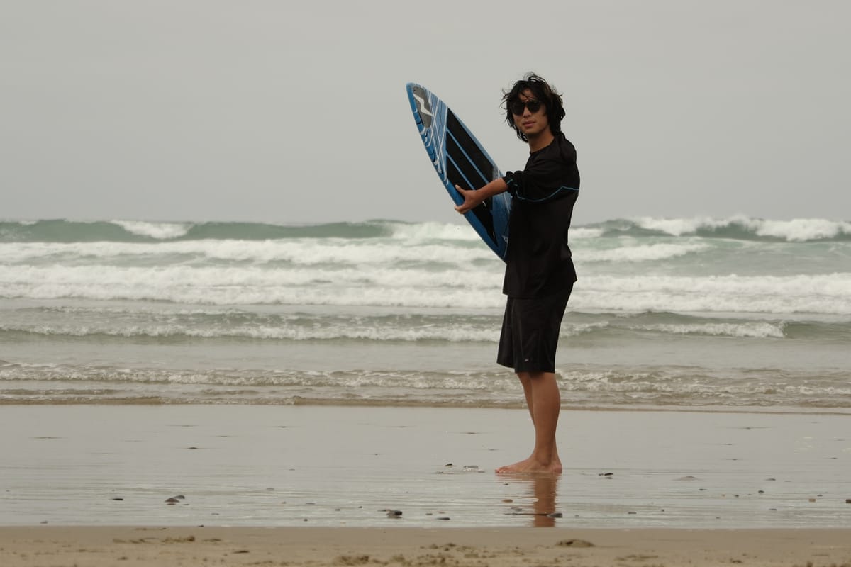 Gabe Lusk on a grey beach, holding a surfboard.